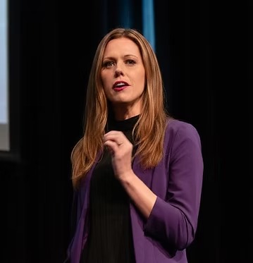 Rebecca Maklad, waist-up portrait in a purple blazer, gesturing mid-speech against a dark background
